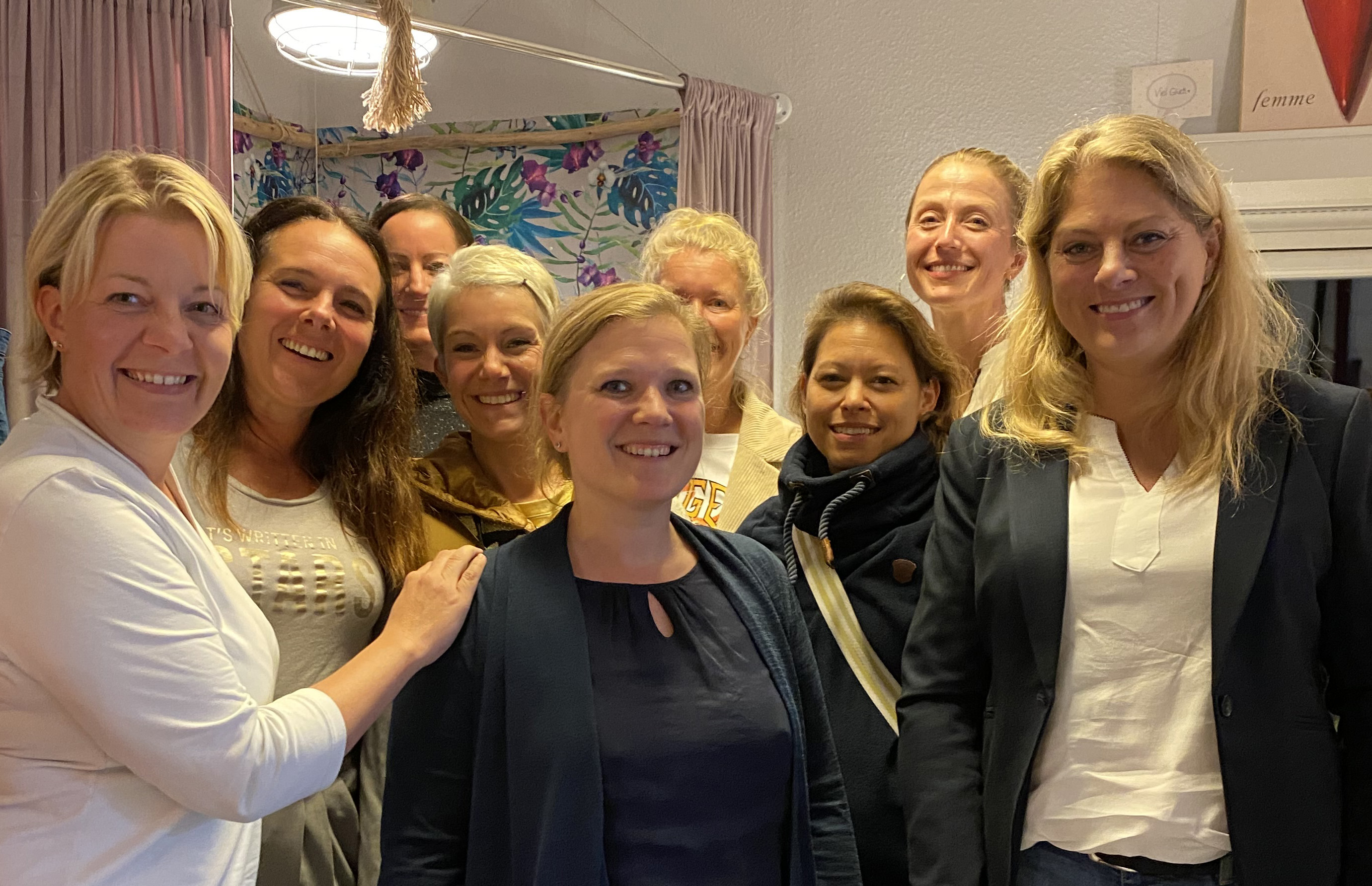 Neun Frauen stehen in einer Boutique und lachen.